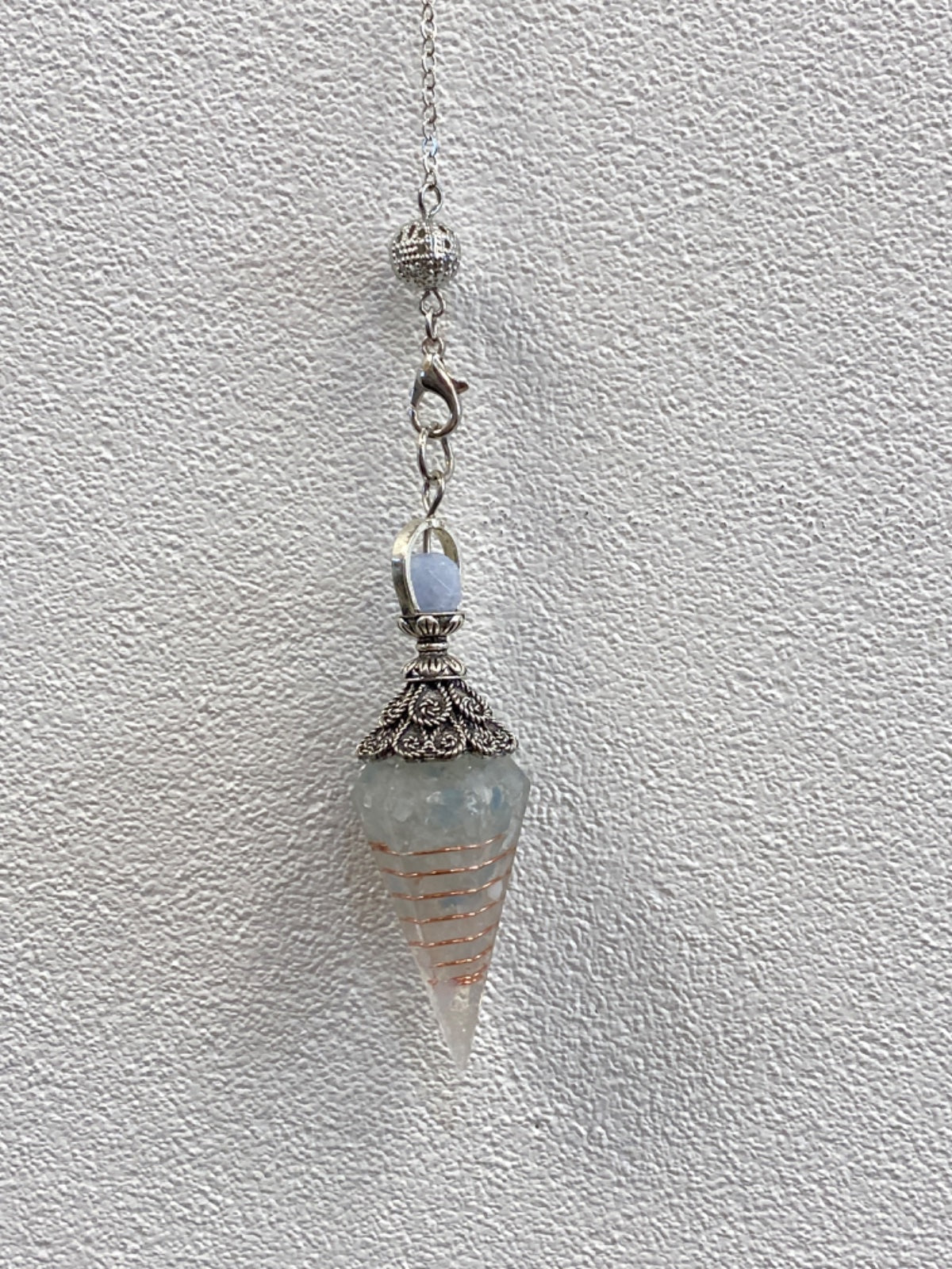 Celestite crystal resin pendulum hanging from a silver chain against a white textured wall.