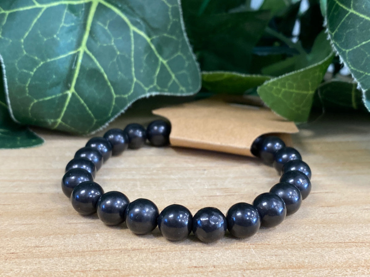 Black shungite bead bracelet with 8mm round polished beads, displayed on light wood with green leaves behind.
