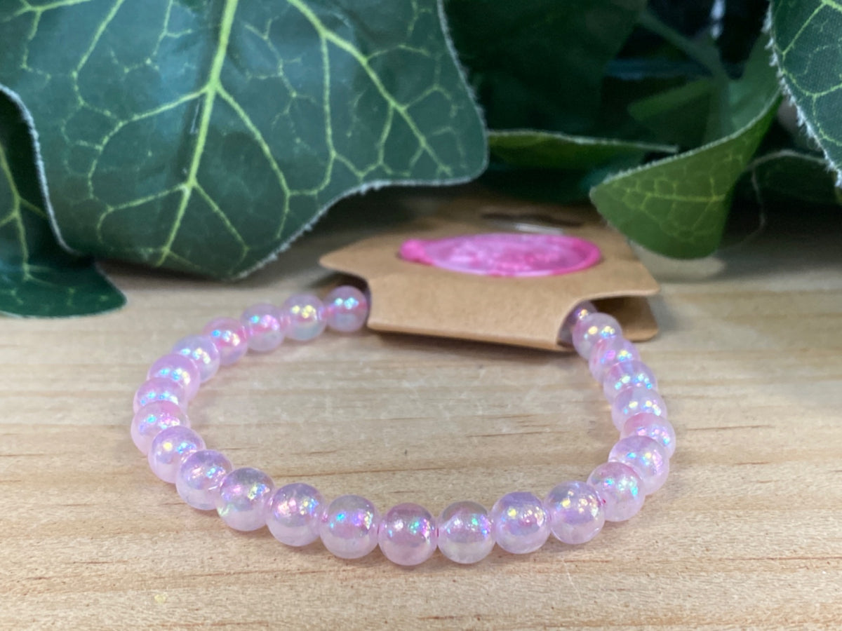 Aura Rose Quartz 6mm bead bracelet displayed on wooden surface with green foliage and kraft packaging in background.