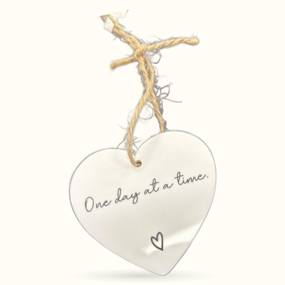 White heart-shaped plaque with 'One day at a time' text, hung from twine and driftwood on white background