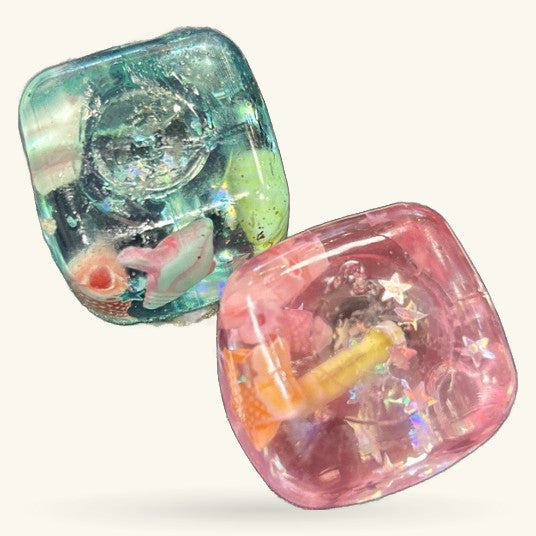 Two square squishy toys with clear gel filling containing miniature ocean-themed objects in teal and pink colours