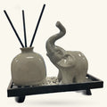 Gray ceramic elephant reed diffuser with three black sticks in round vase, on black tray with pebbles