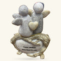 Two grey ceramic angel figures with wings and a golden heart, sitting on a carved stone base engraved with 'Love makes two hearts one'
