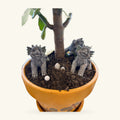 Terracotta pot with two grey dragon figurines, a live plant with green leaves, and small white stones in dark soil.