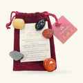 Five polished tumble stones in shades of gold, black, rust, and grey, displayed with a cream information card in a burgundy velvet drawstring pouch.