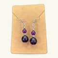 Purple bead earrings with dark round stones and silver-toned metal hooks, displayed on a kraft card.