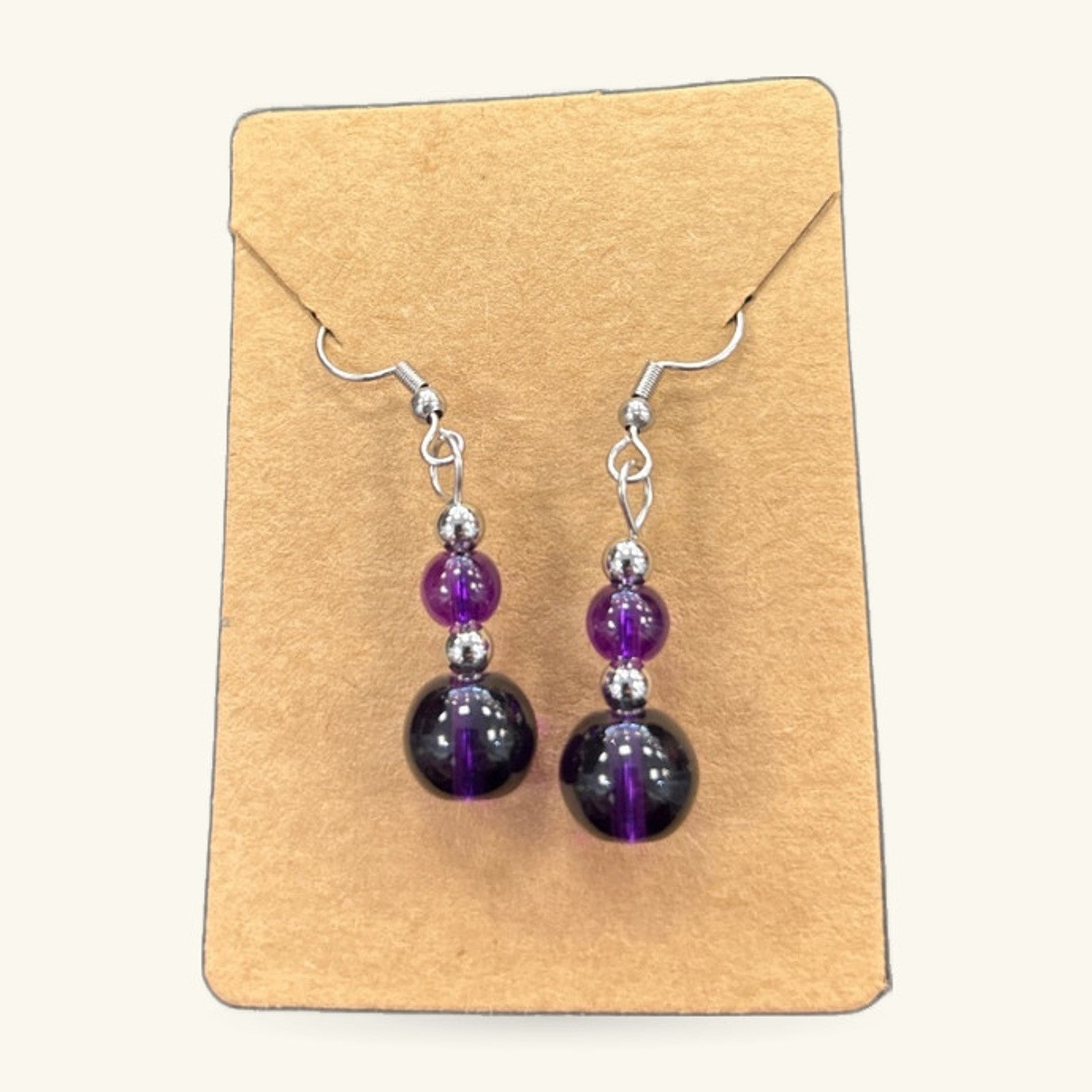 Purple bead earrings with dark round stones and silver-toned metal hooks, displayed on a kraft card.