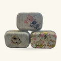 Three vintage-style metal pill boxes stacked in a triangle, each decorated with different floral patterns in soft colours.