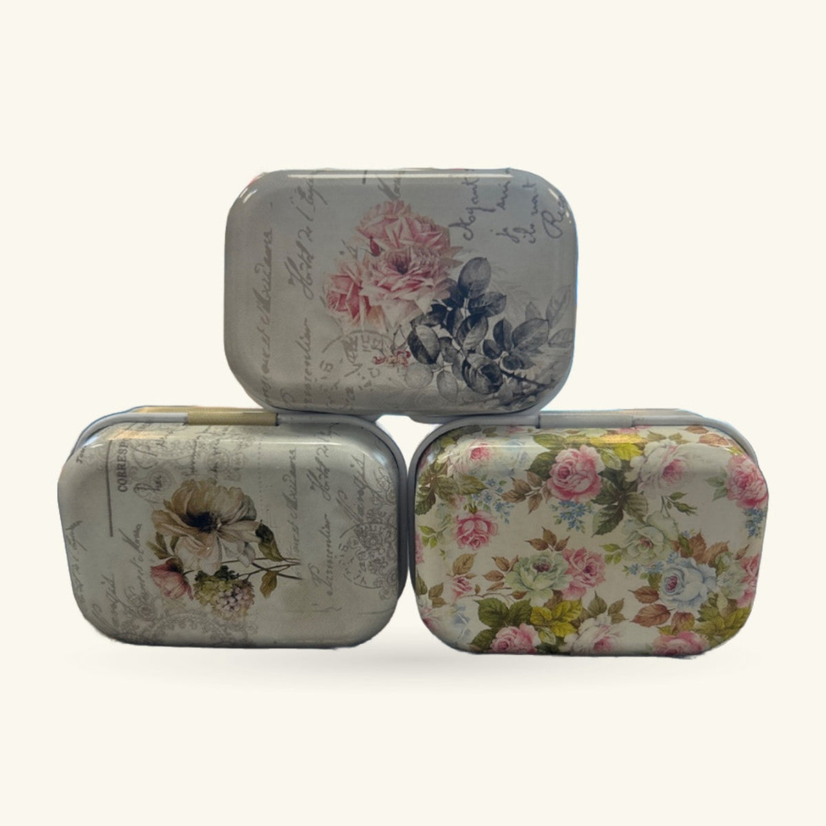 Three vintage-style metal pill boxes stacked in a triangle, each decorated with different floral patterns in soft colours.