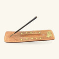 Wooden incense holder with brass stars and circles, black incense stick resting in groove