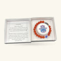 Hamsa Evil Eye Carnelian Bracelet with red-orange beads, blue enamel eye bead, and Hamsa charm, displayed in open white gift box with information card